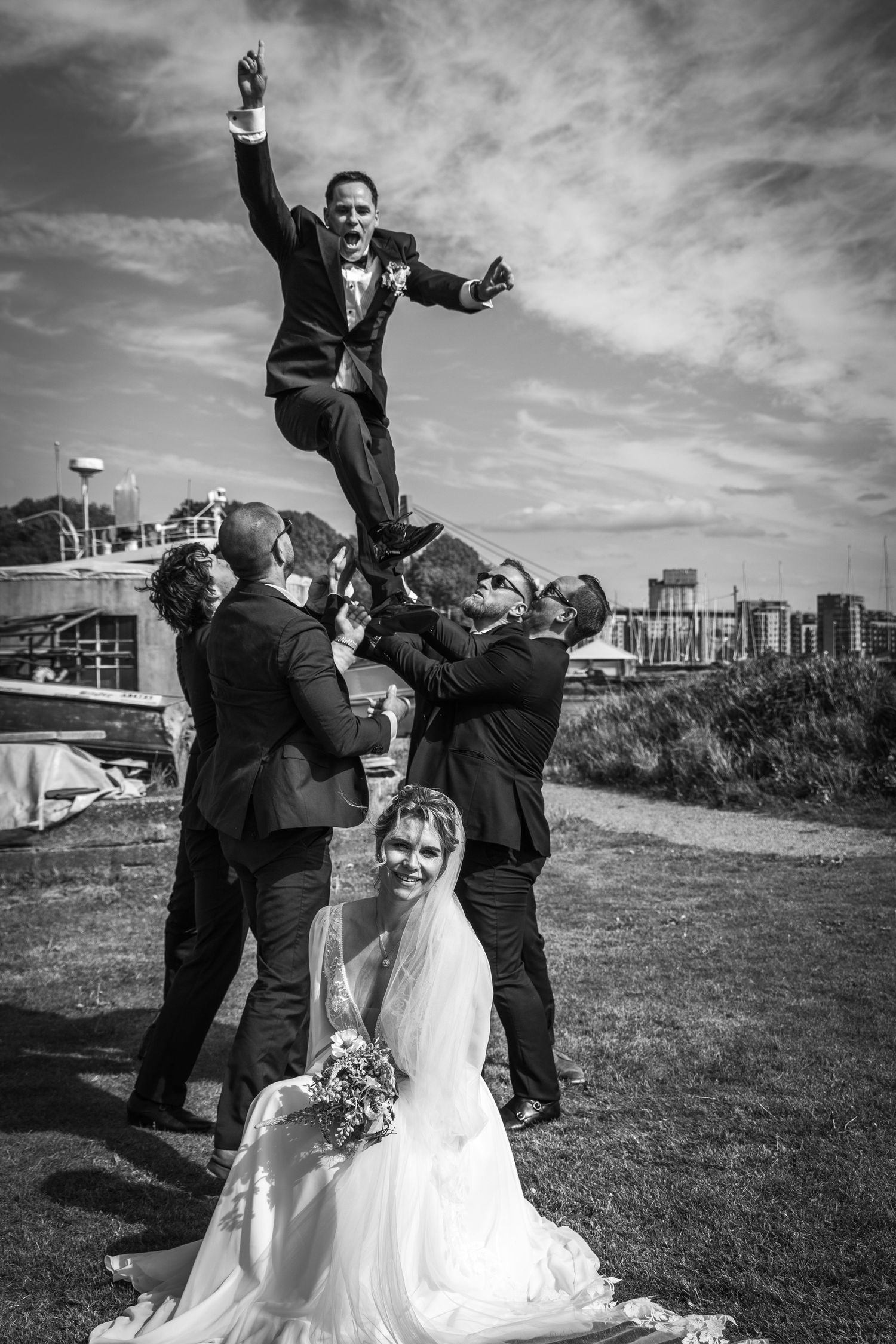 Black and white photo of groomsmen lifting groom overhead while bride smiles in foreground.
