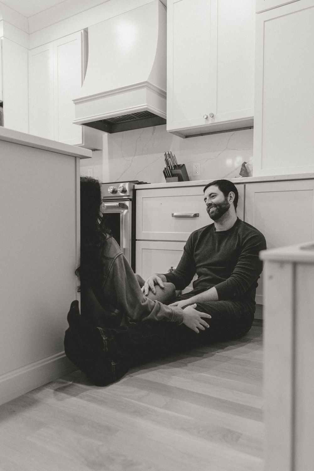 Two people sit casually on a kitchen floor having a relaxed conversation in a modern white kitchen.