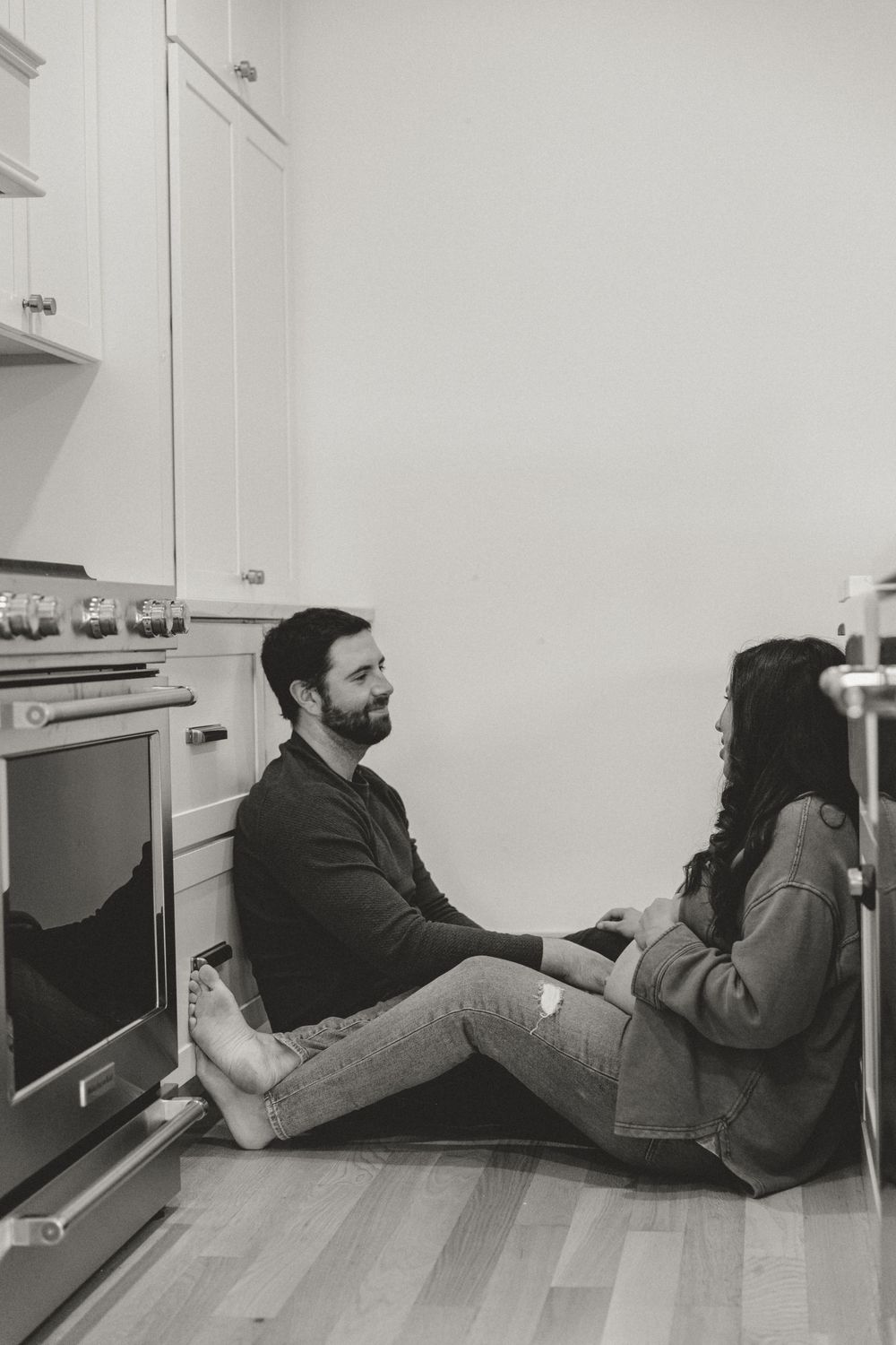A casual lifestyle photo of two people sitting on hardwood floors in a modern white kitchen.