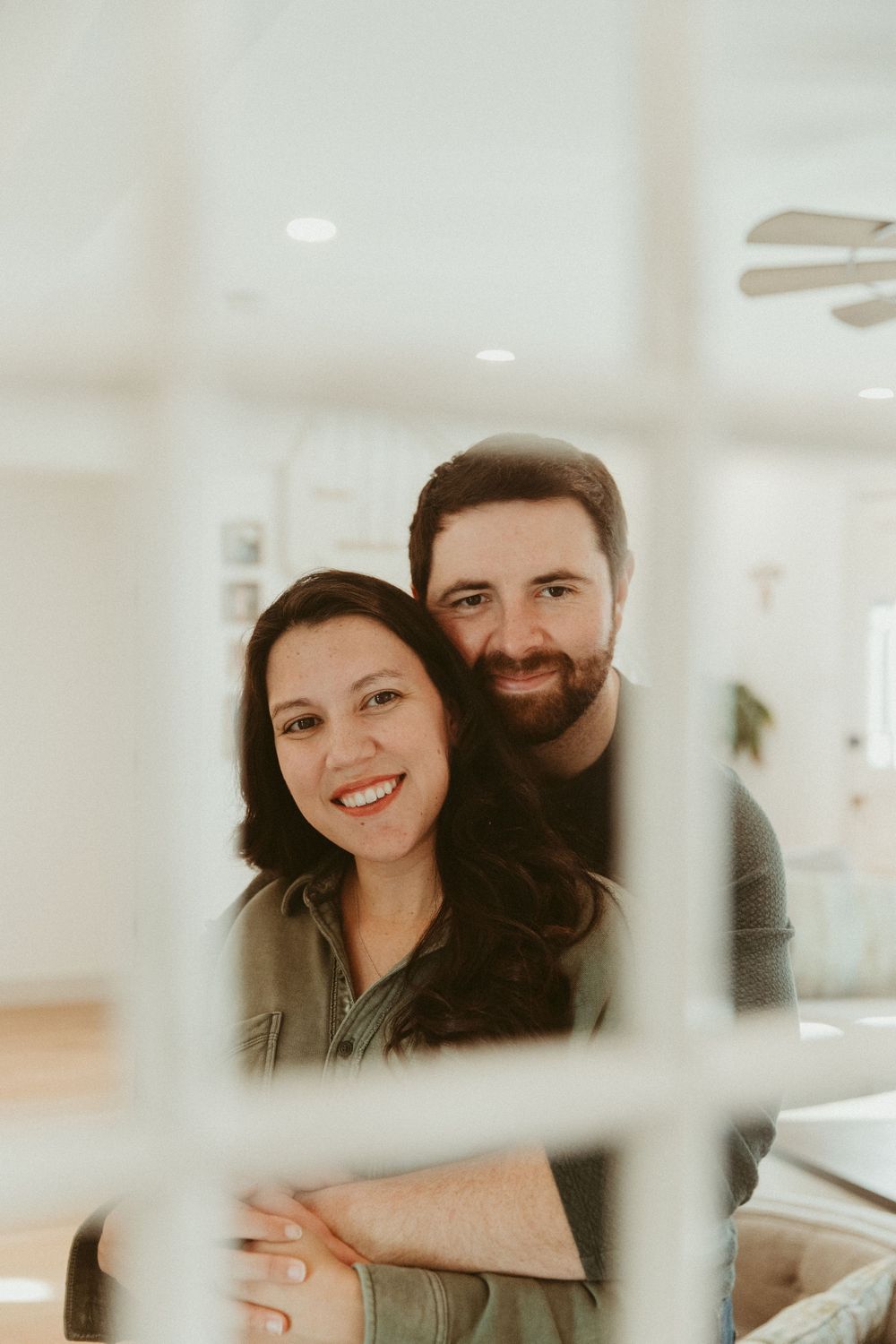 A happy couple poses for a portrait through window panes in a bright interior setting.