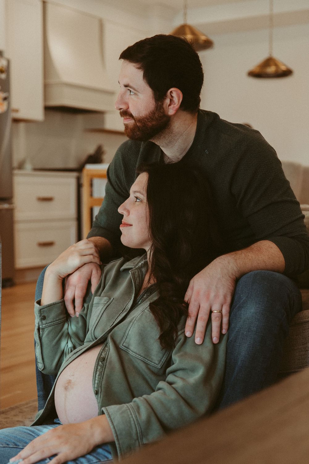 Cozy maternity photo of couple sitting on grey sofa in warm-lit kitchen.