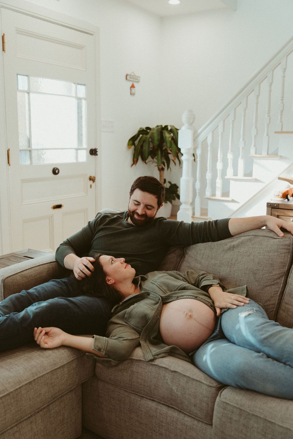 Candid maternity photo showing couple laughing while relaxing on grey sofa.