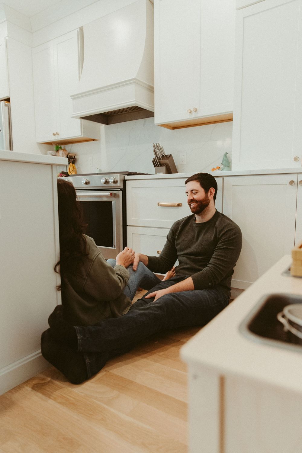 A couple relaxes on the kitchen floor, sharing a casual moment in their modern white kitchen.