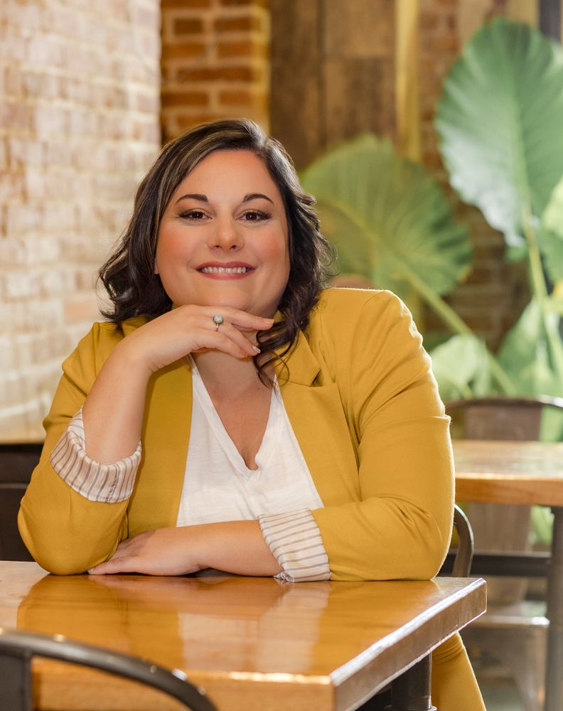Portrait of a person in a mustard yellow blazer sitting at a wooden table with tropical decor.