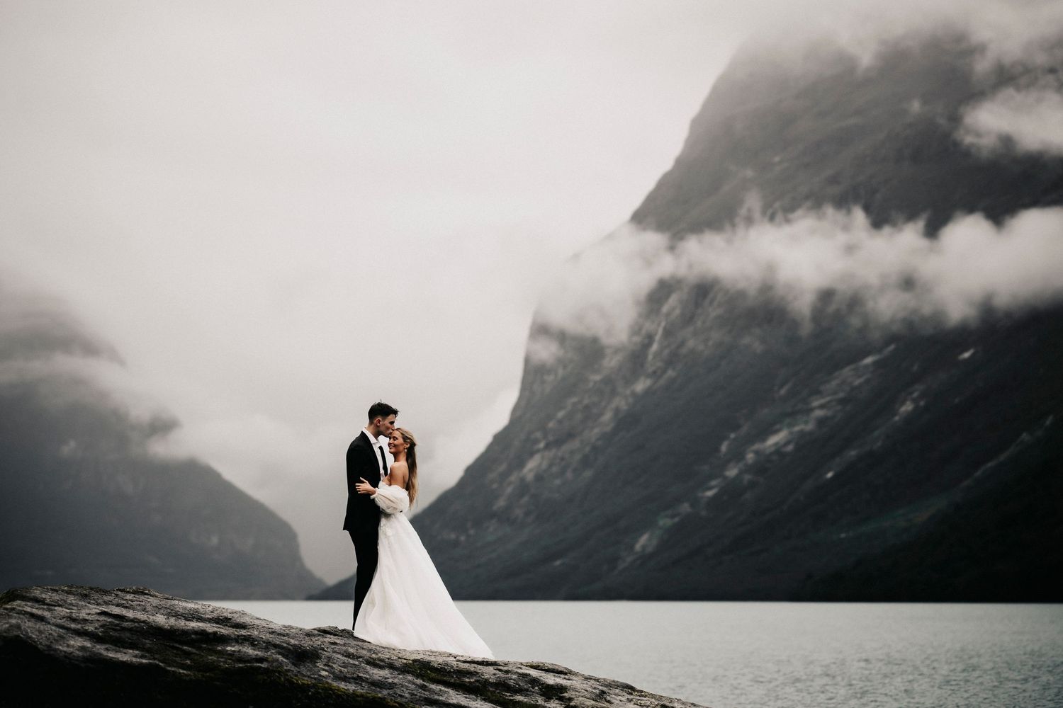 Silhouettes embrace on rocky outcrop overlooking misty mountain lake with dramatic peaks shrouded in clouds.