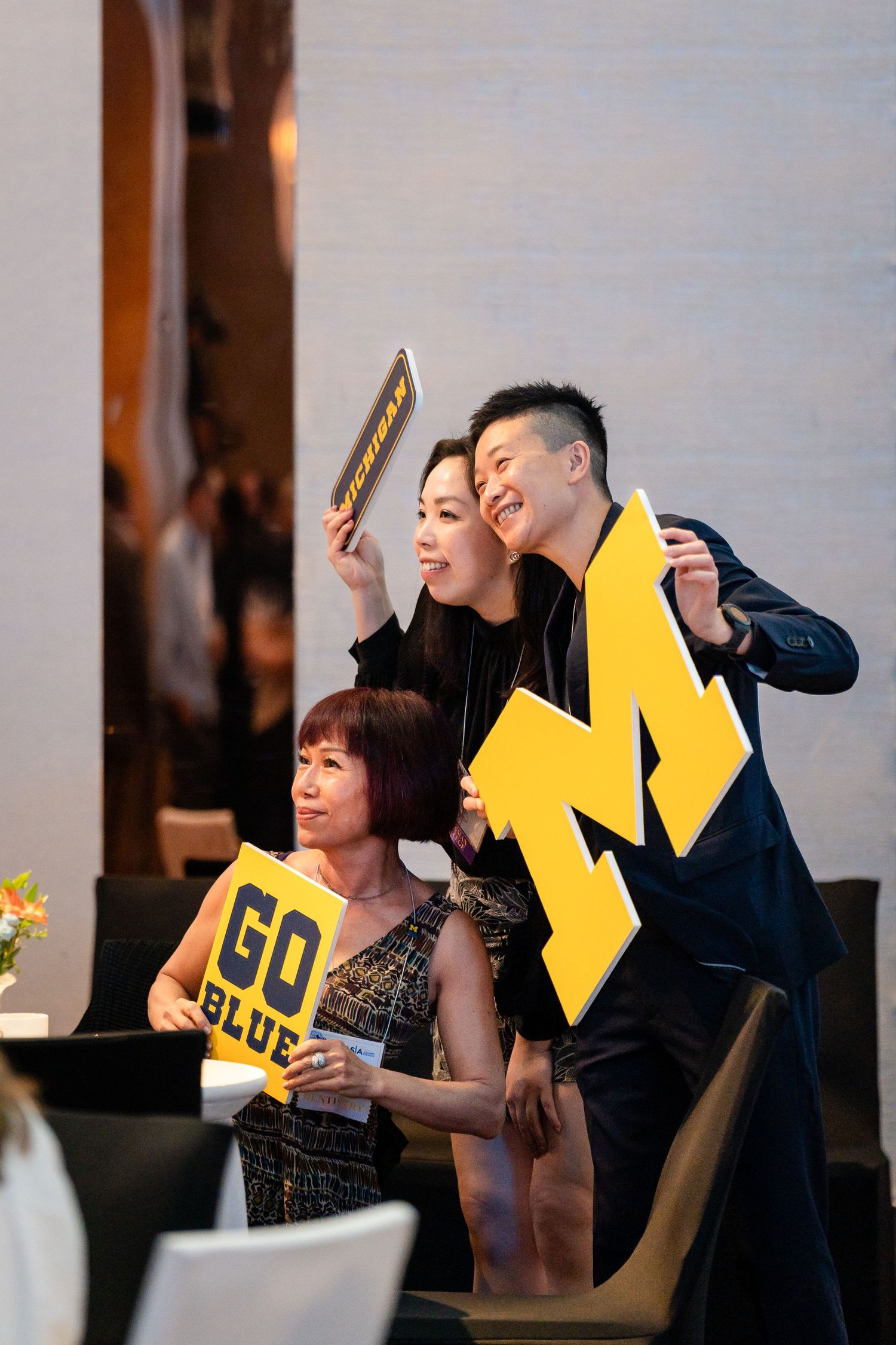 Hands with multicolored nails hold a white canvas tote bag featuring a yellow M and 'Alumni' text in blue letters.