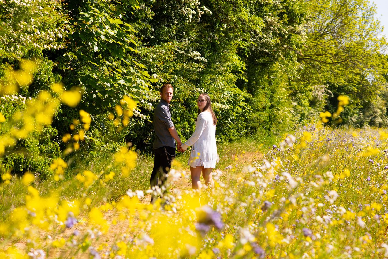 Two silhouettes stand together in a sunny wildflower meadow filled with yellow and white blooms on a summer day.