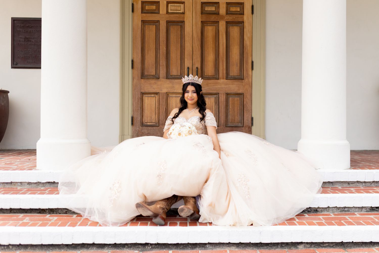 A bride in a flowing champagne-colored gown and flower crown sits gracefully on white stone steps by wooden doors.