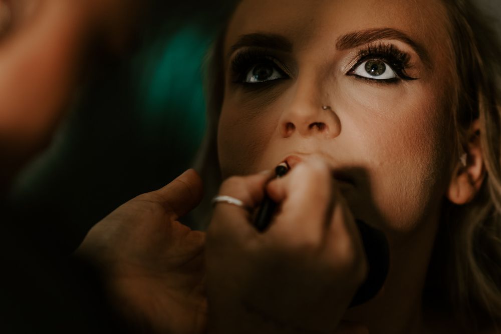 Closeup shot of makeup application on lips with dramatic eye makeup visible in moody lighting.