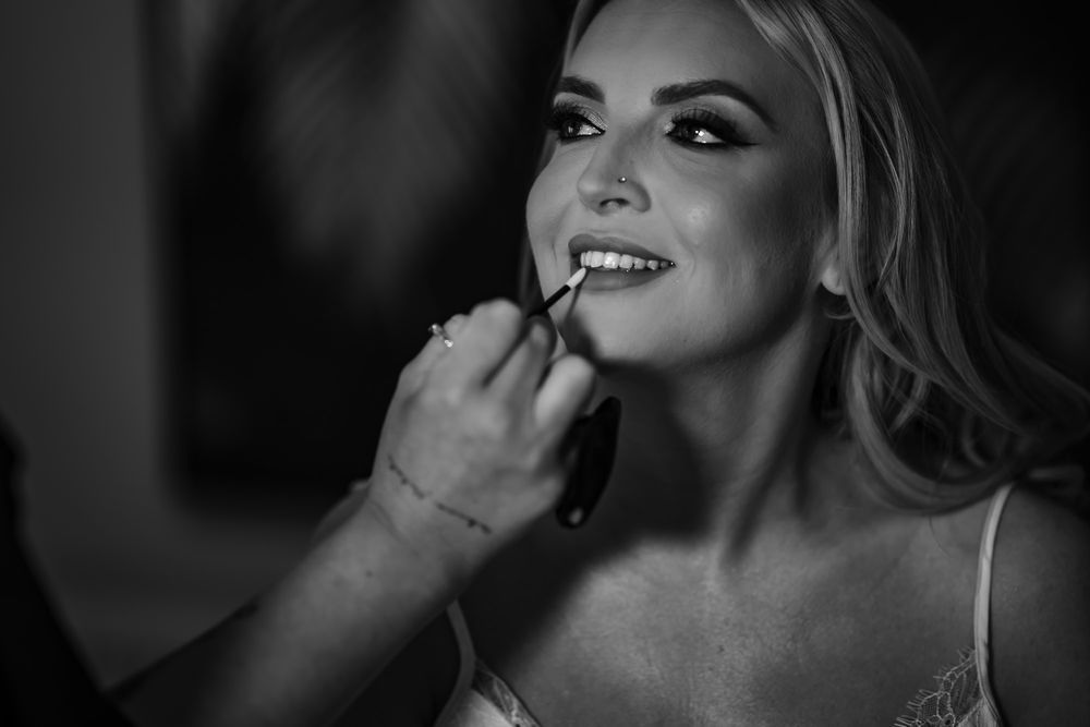 Black and white photo shows makeup artist applying lipstick during bridal beauty preparation session.