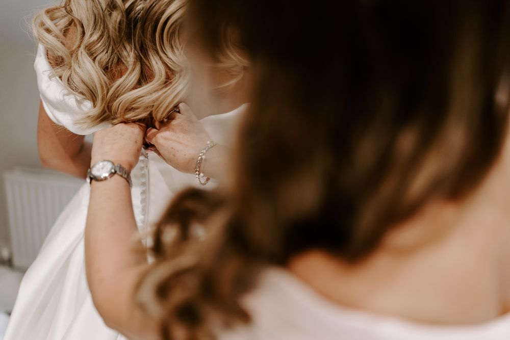 A close-up detail shot of hands adjusting a white wedding dress with curled hair visible in the background.