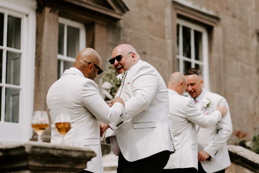 Three groomsmen in white jackets share a candid laughing moment during a wedding celebration.