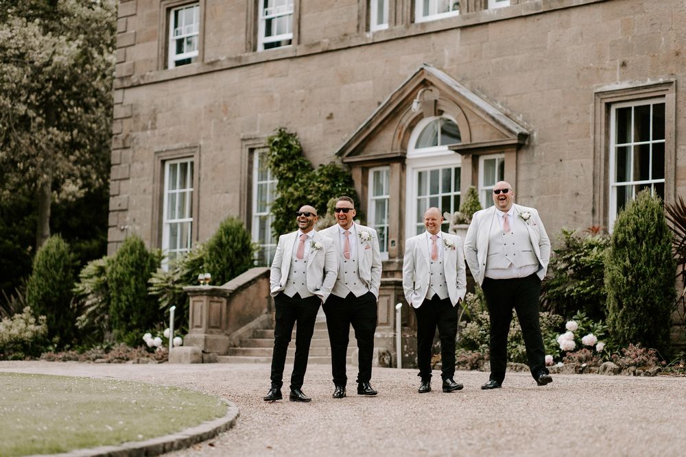 A group in matching white dress shirts and dark pants pose together outside an elegant stone manor house.