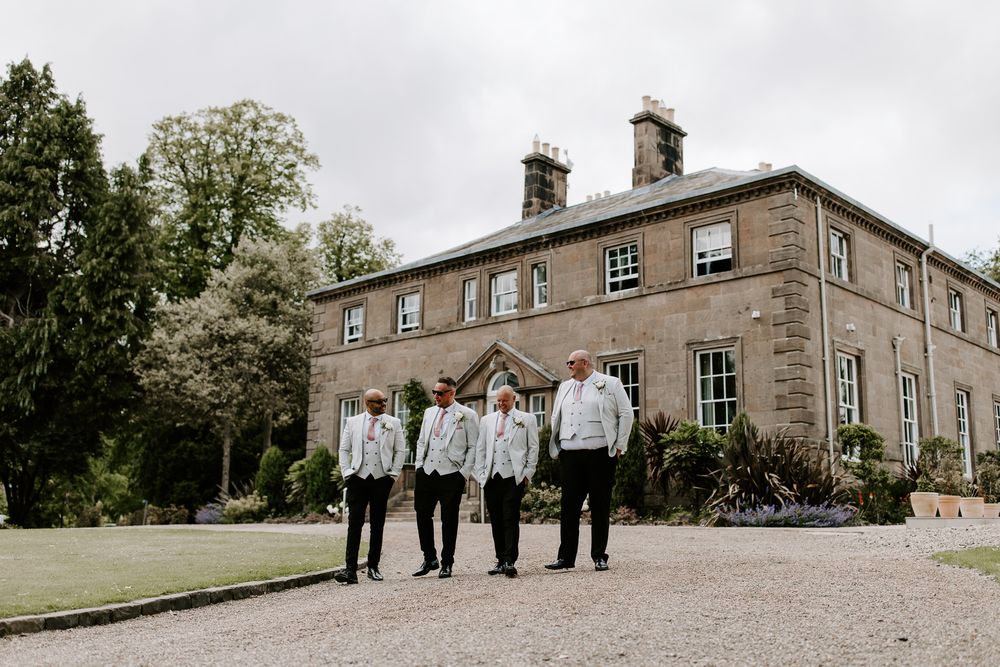 Wedding party poses on manicured lawn in front of elegant stone Georgian mansion on overcast day.