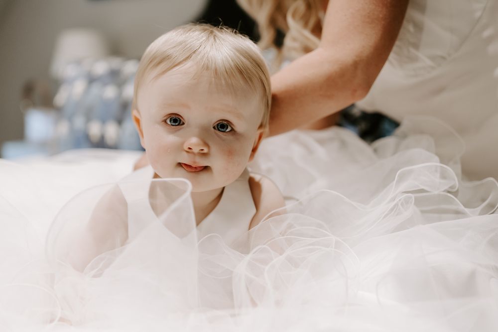 A baby in a white tulle dress smiles while sitting amidst soft, fluffy white fabric and tulle.