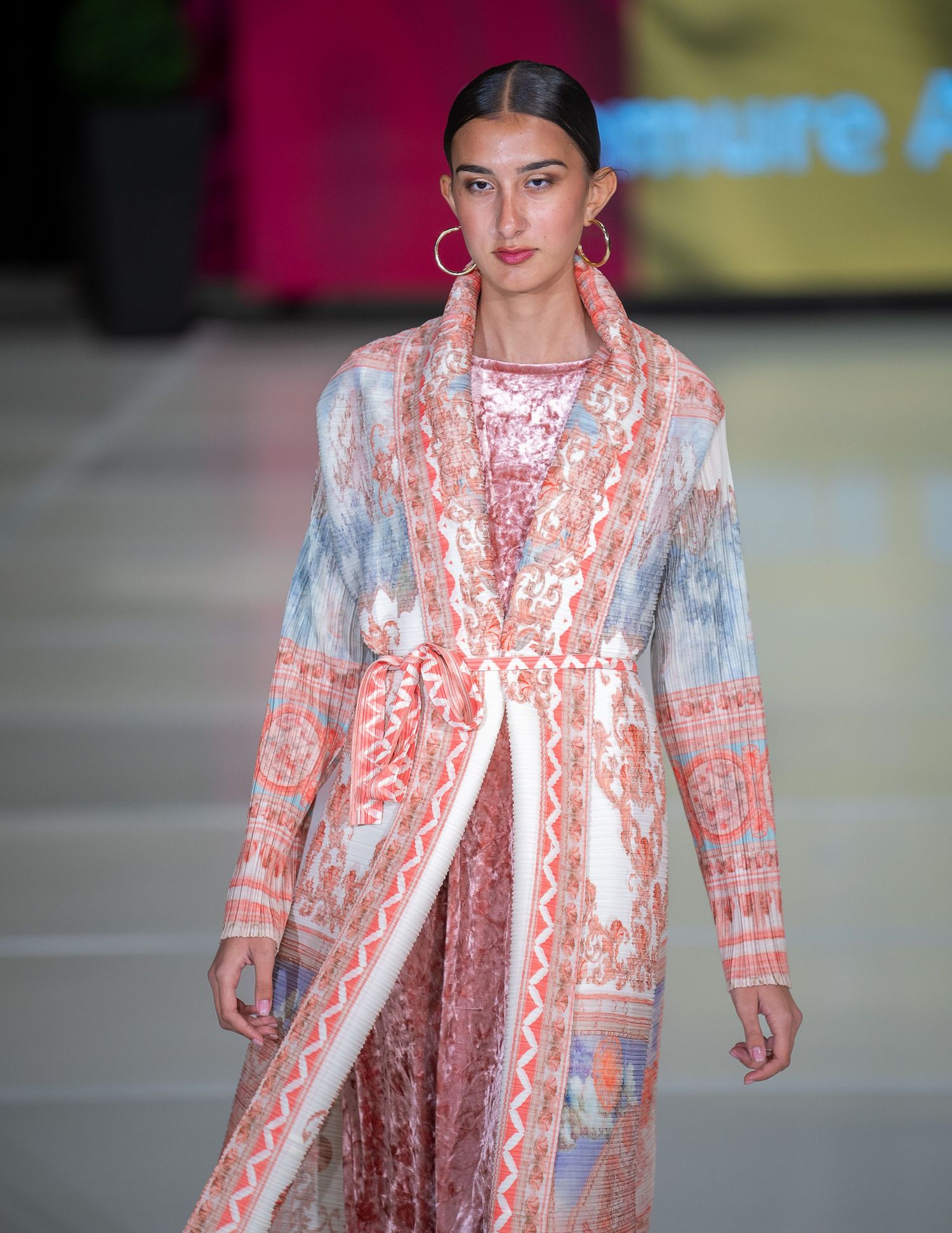Model walks runway wearing pink and blue patterned long coat with ethnic-inspired design at fashion show.