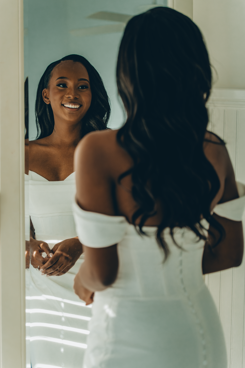 A reflection in a mirror shows an elegant off-shoulder white dress with flowing dark hair and a radiant smile.