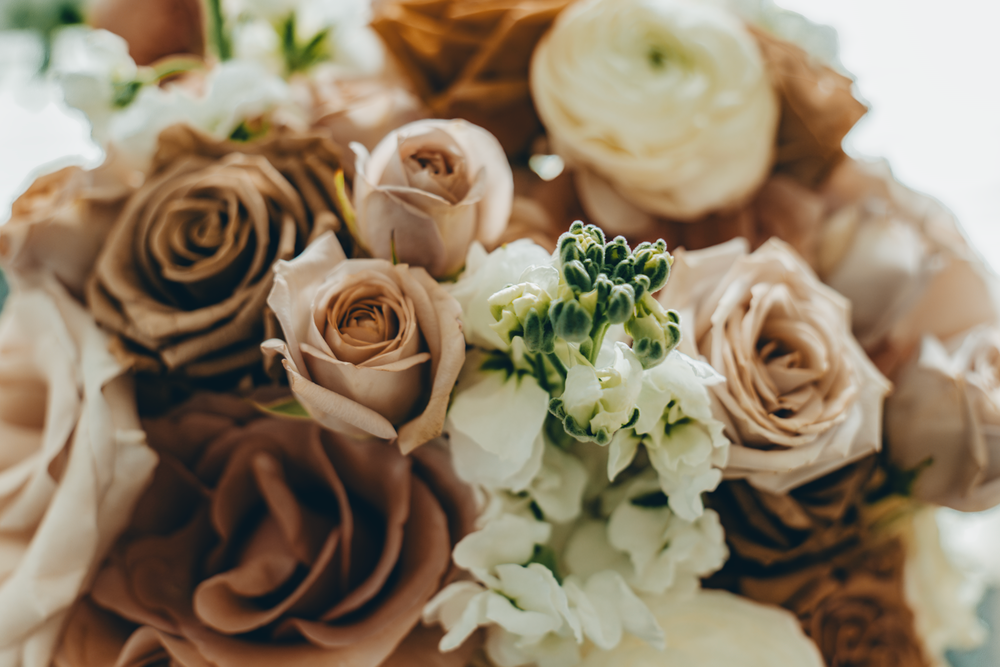 Close-up view of an elegant wedding bouquet featuring mocha-colored roses and white ranunculus flowers in soft focus.