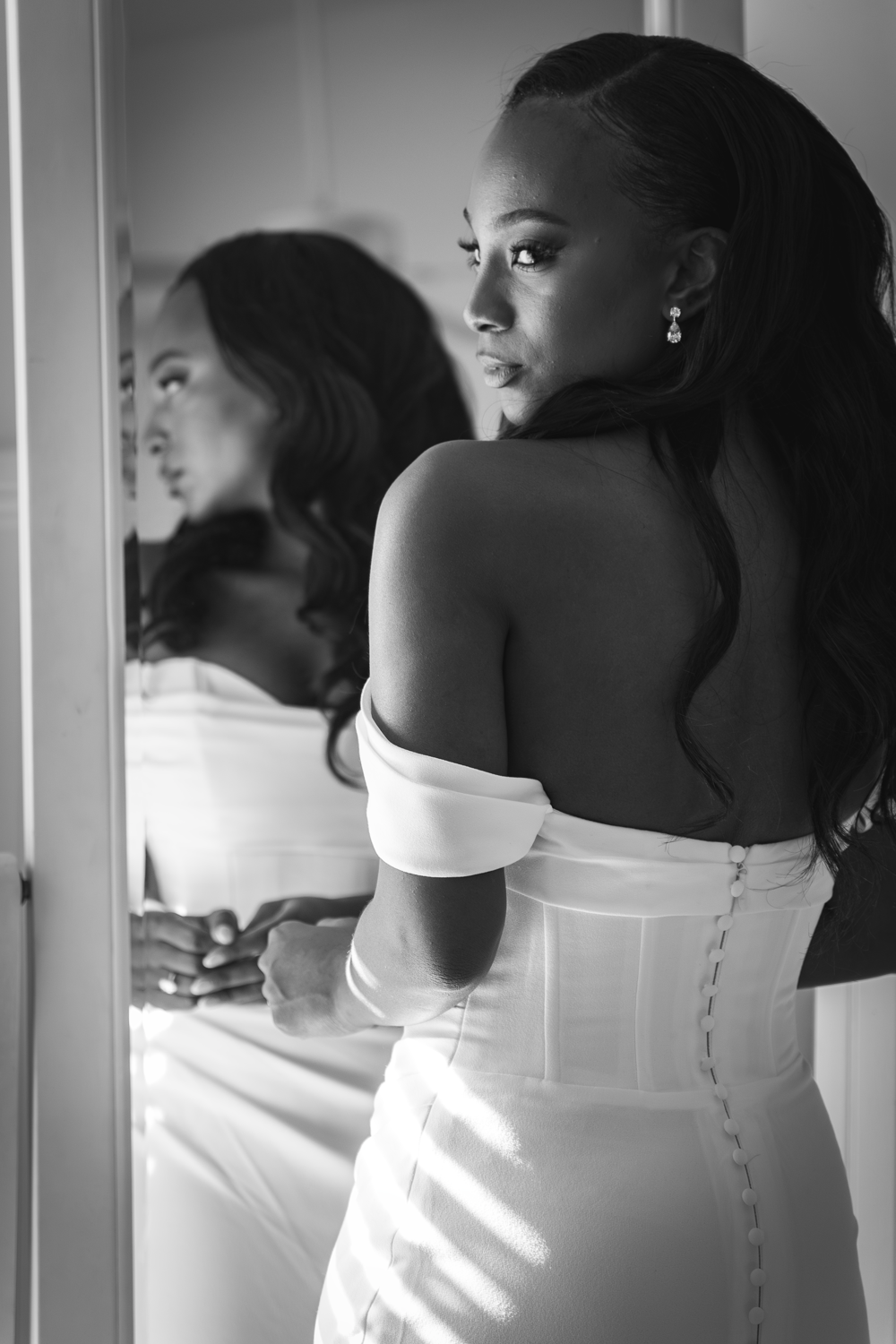 Black and white portrait of a person in an off-shoulder white dress looking over their shoulder near a mirror.