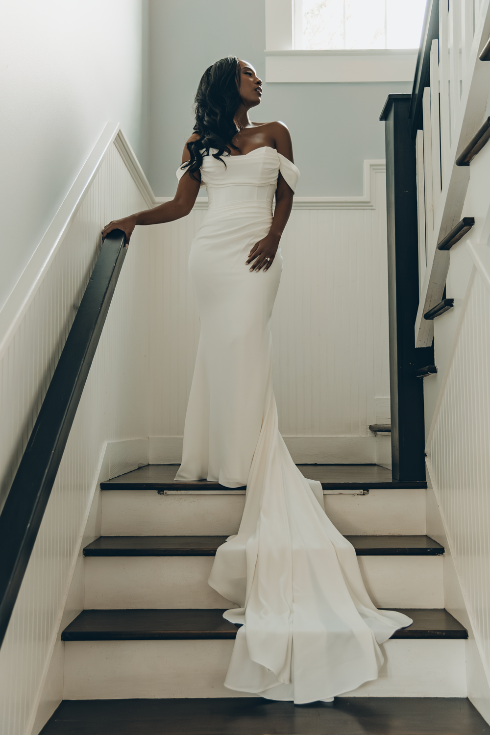 A bride in an elegant off-shoulder white mermaid wedding gown poses gracefully on a staircase with black steps and white walls.