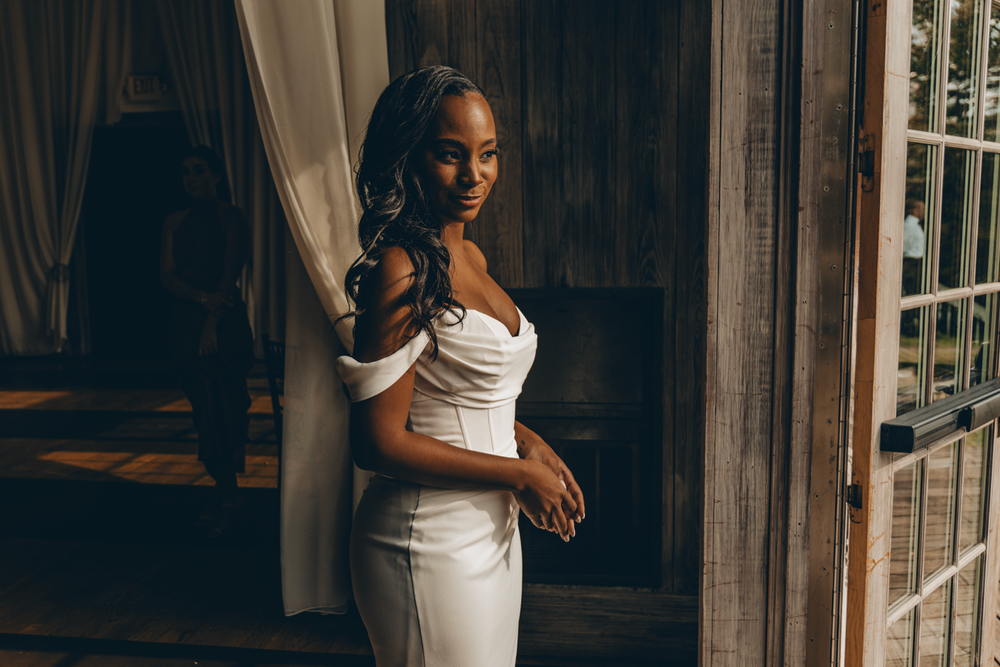 Elegant figure stands by window in flowing white off-shoulder dress with romantic golden hour lighting.