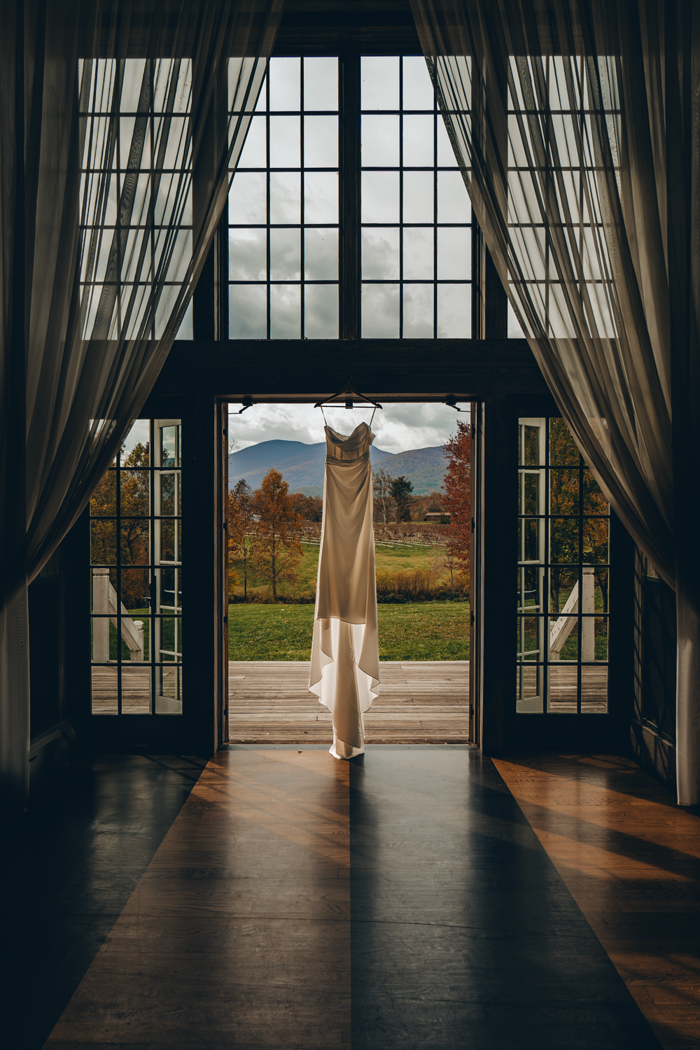 A white wedding dress hangs in elegant French doors overlooking autumn mountains and foliage on a moody overcast day.