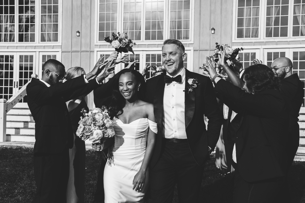 A joyful wedding party celebrates outside a building with large windows as a couple holds their hands up triumphantly.