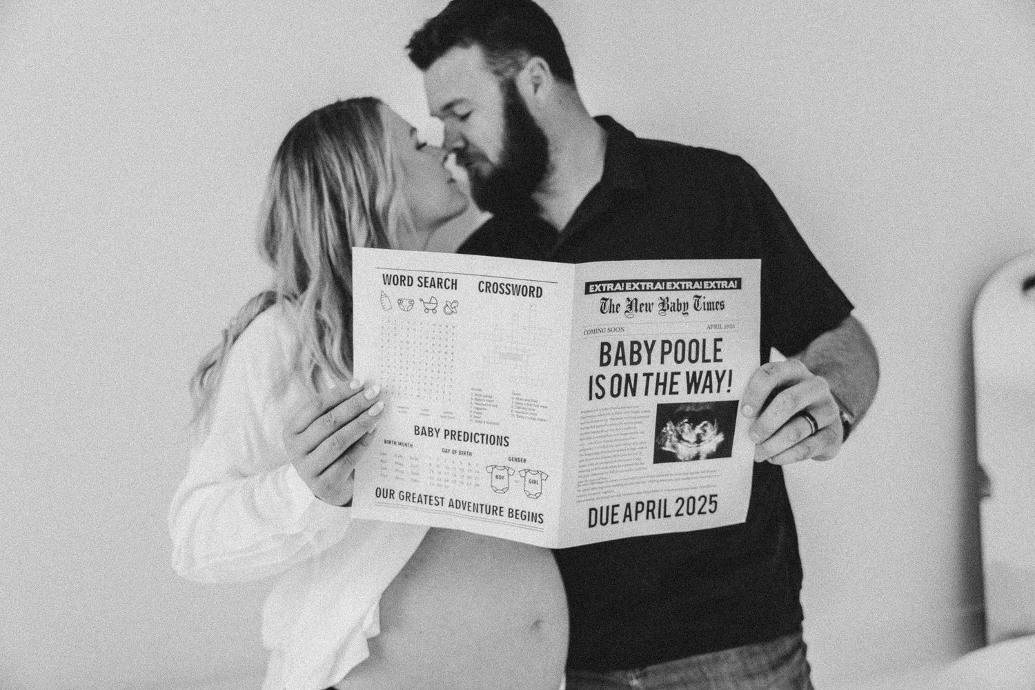 Couple shares intimate moment while holding birth certificate style pregnancy announcement against white wall background.