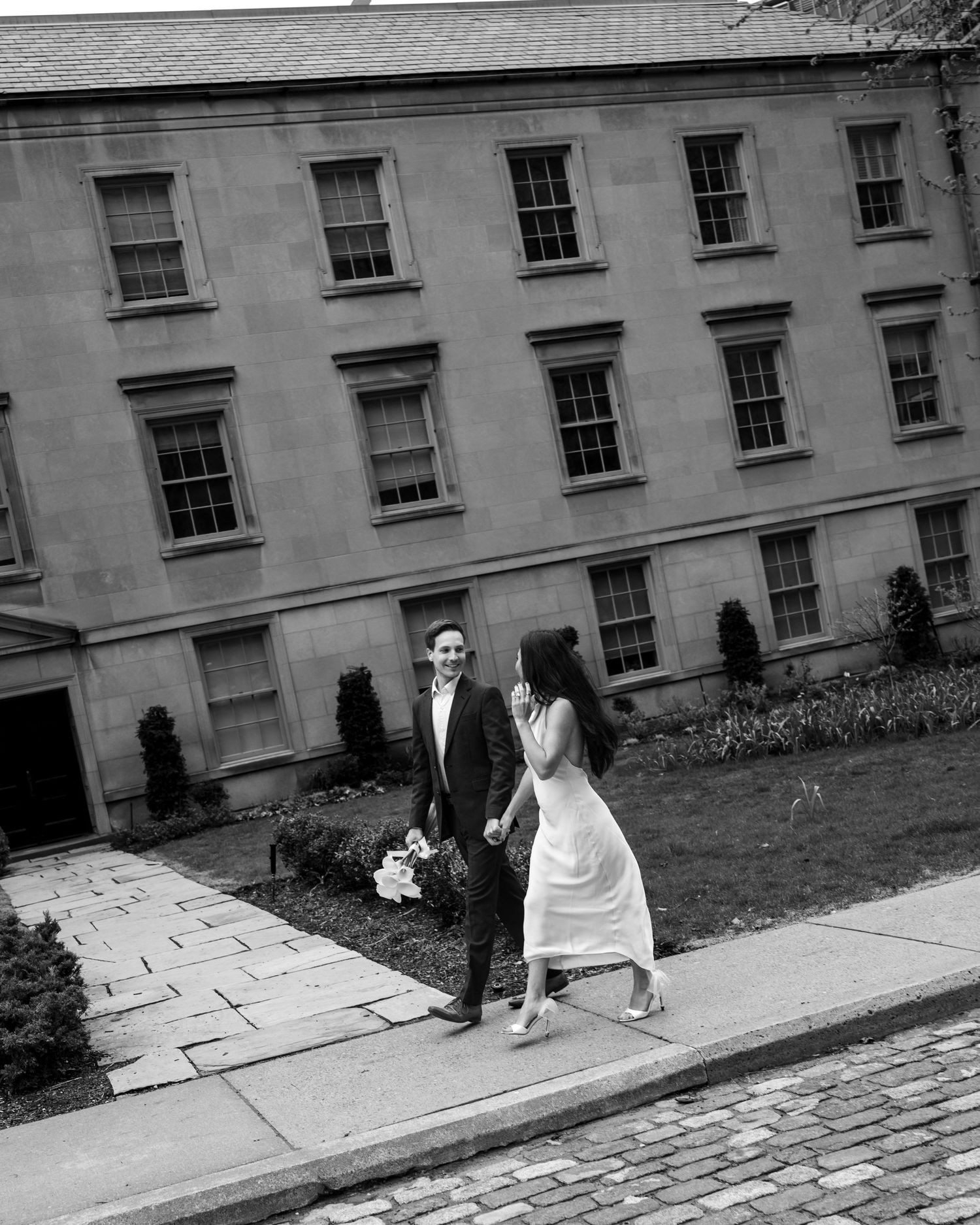 A couple stands outside a historic building on a cobblestone path in black and white photography.