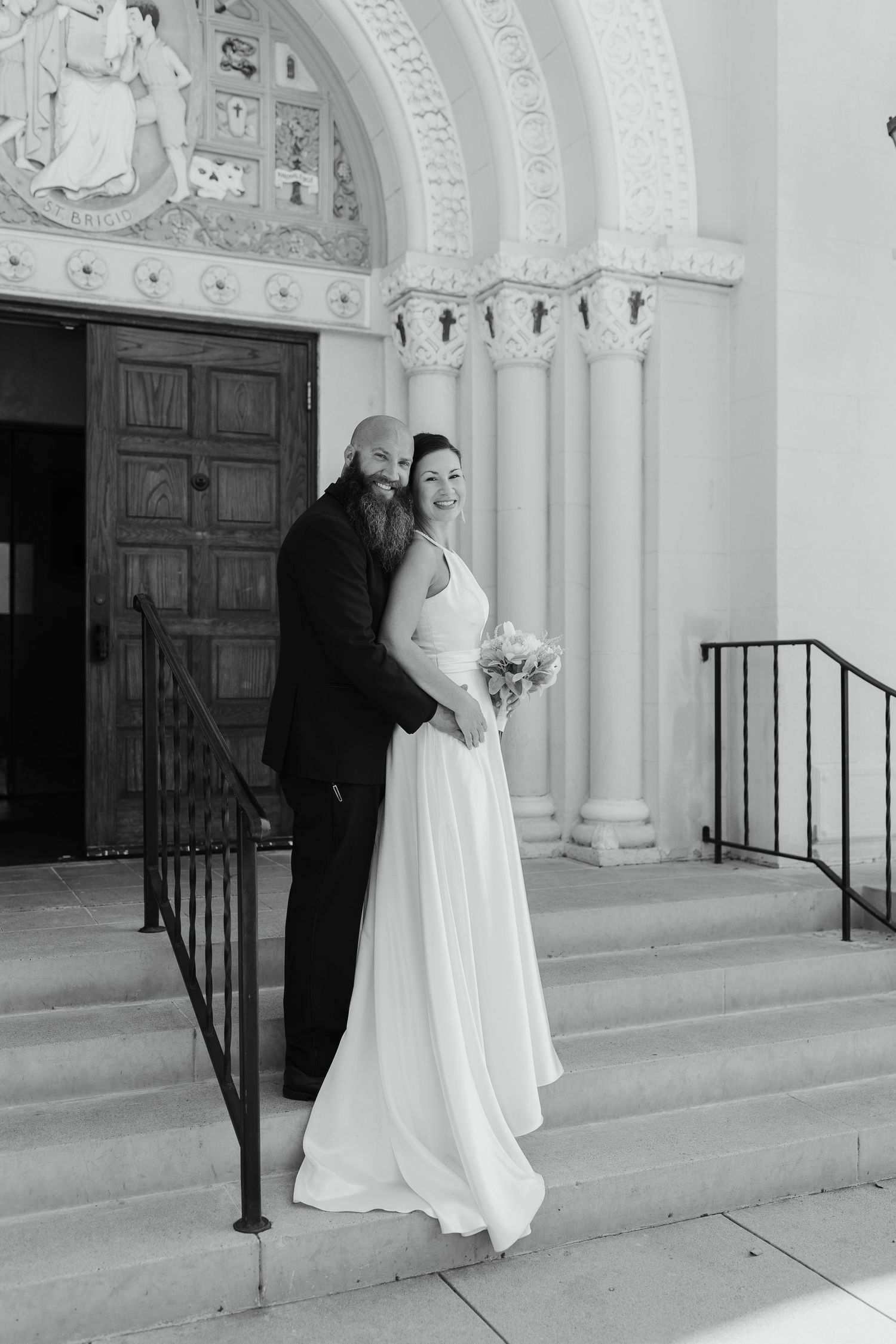 Couple in wedding attire standing on stone steps outside an ornate church building, embracing and smiling