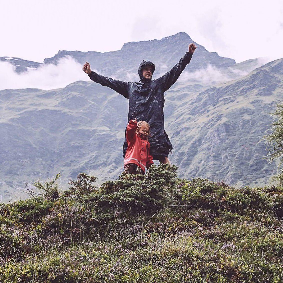 Enthusiastic hiker celebrates on mountain summit against backdrop of snow-capped peaks.
