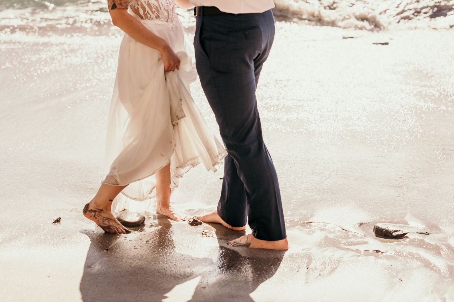 Two pairs of feet stand close together on wet sand at the beach as gentle waves wash over them.