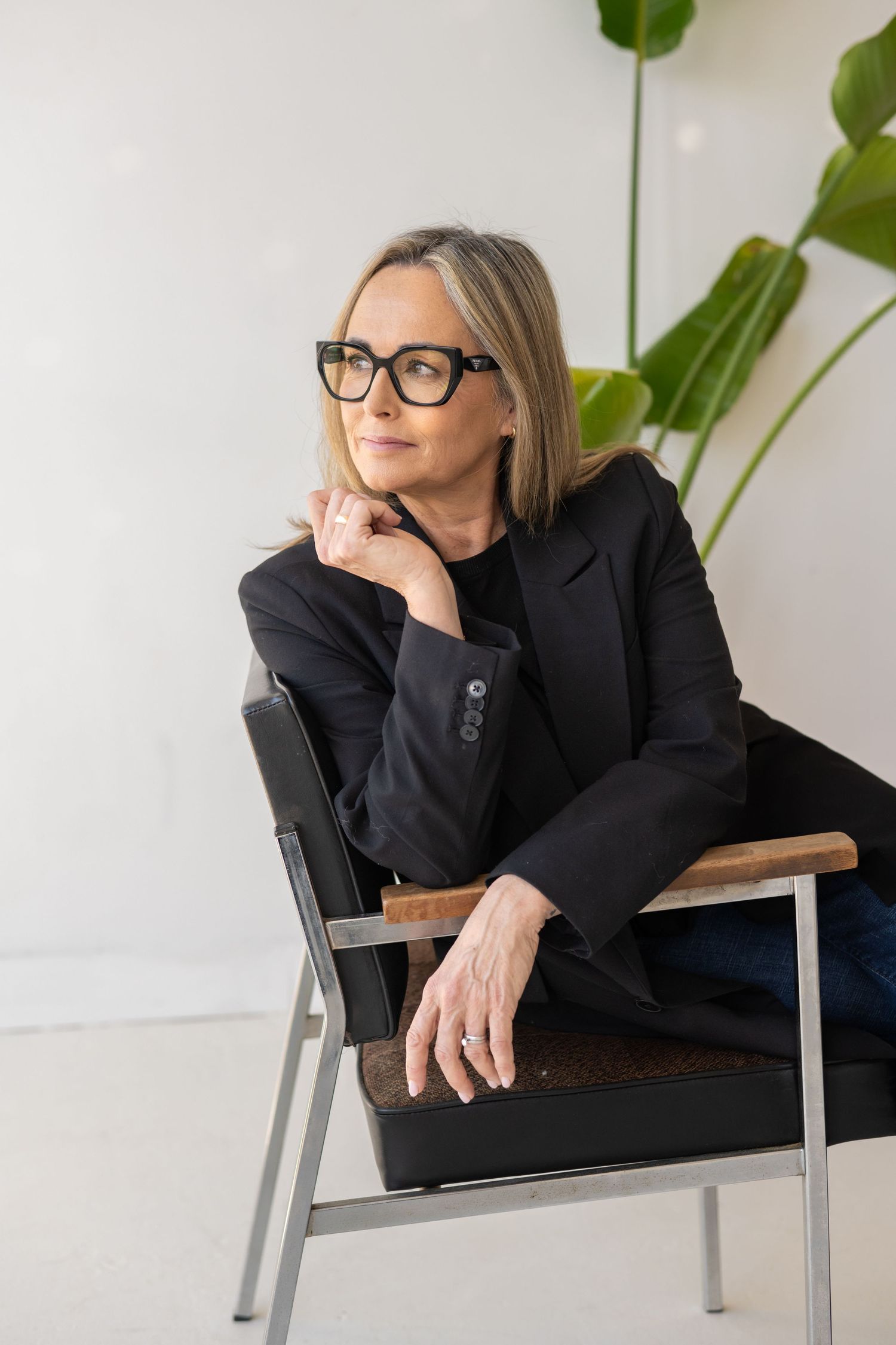 Professional wearing black blazer and glasses sits thoughtfully in modern chair with houseplant in background.