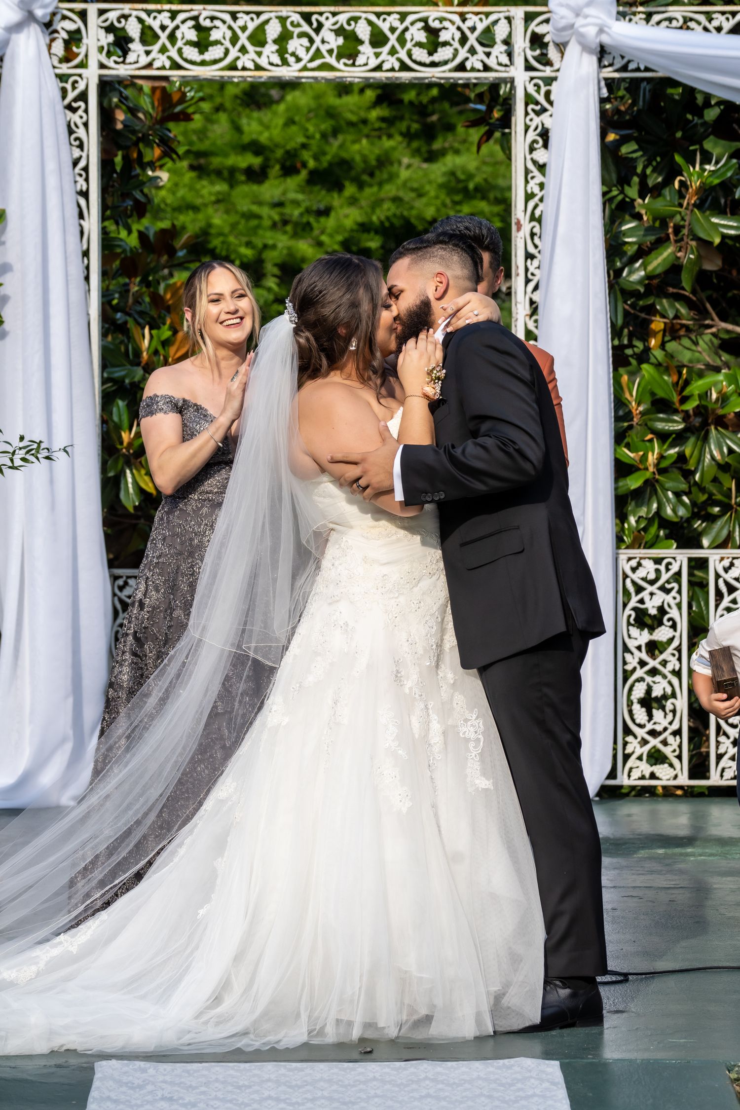 A wedding ceremony kiss captured under an ornate white gazebo with flowing fabric decorations.