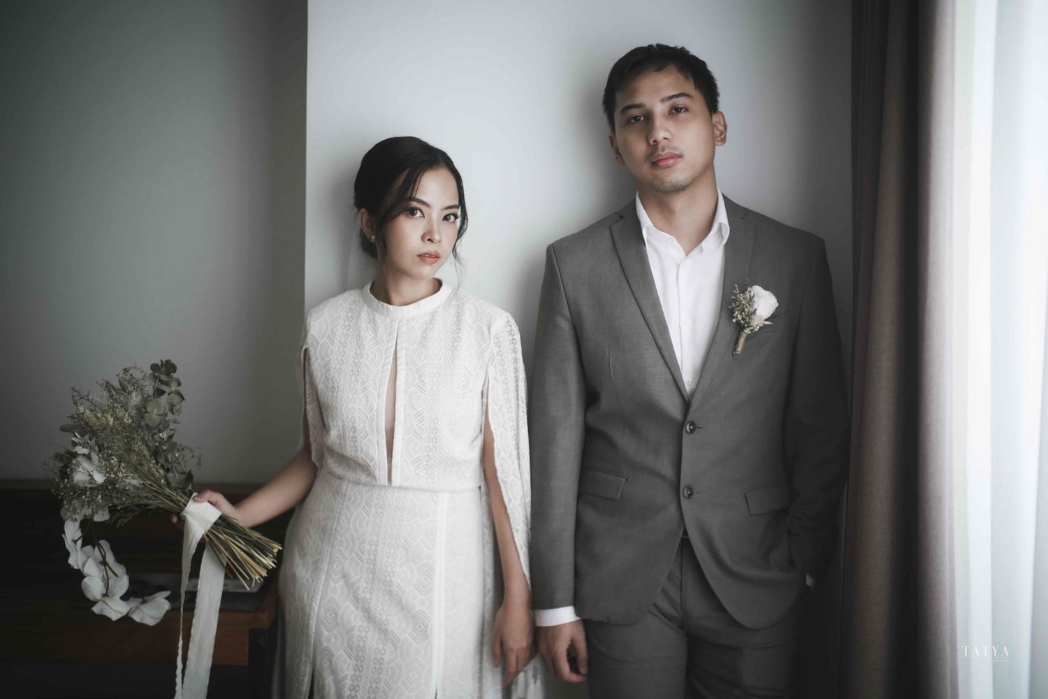 Pasangan pengantin berdiri berdampingan di dalam ruangan dengan pencahayaan alami, memancarkan kesan tenang dan elegan