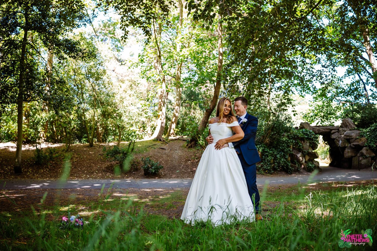 A wedding couple embraces in a sun-dappled garden setting surrounded by lush green trees and natural light in Hesketh Park, Southport