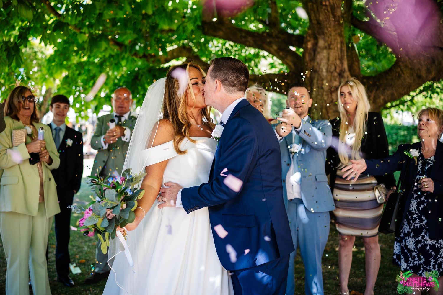Couple shares their first kiss as newlyweds under a leafy tree while wedding guests celebrate with confetti captured by Lynette Matthews