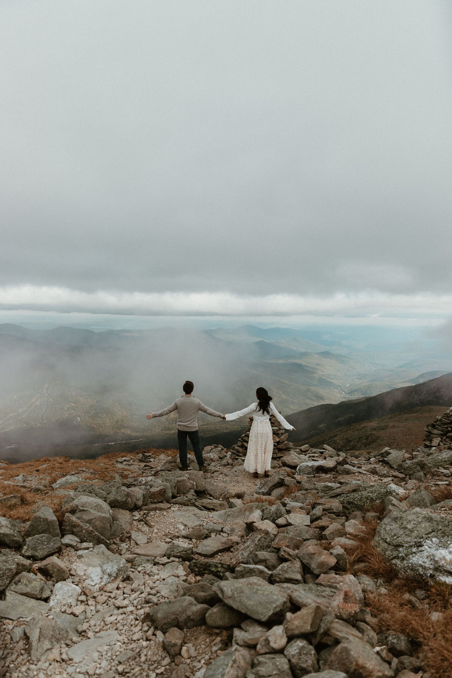 Couple standing on rocky mountain summit overlooking misty valley landscape.