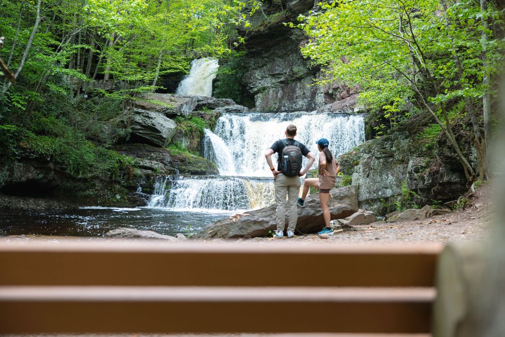 Two hikers stand before a beautiful waterfall in a lush green forest setting.
