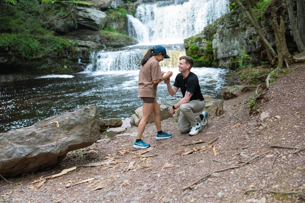 Man kneels on one knee proposing to woman near a scenic waterfall in the woods.