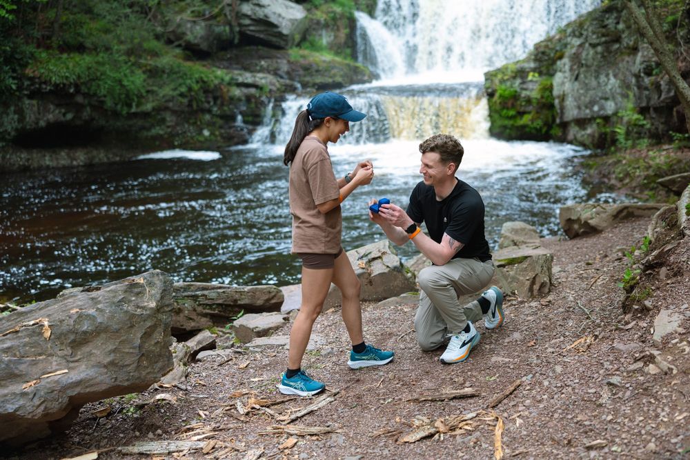 Romantic marriage proposal beside a waterfall as man kneels presenting a ring box.