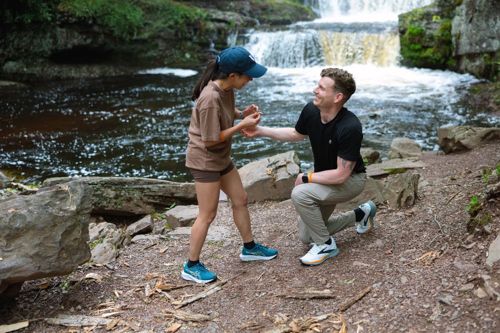 Man on one knee placing engagement ring on womans finger near a forest waterfall.