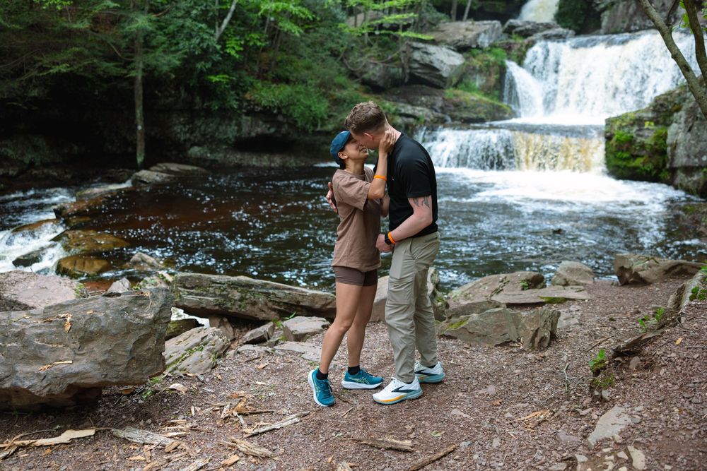 Newly engaged couple shares a kiss beside a picturesque waterfall in the forest. Skytop Lodge engagement.