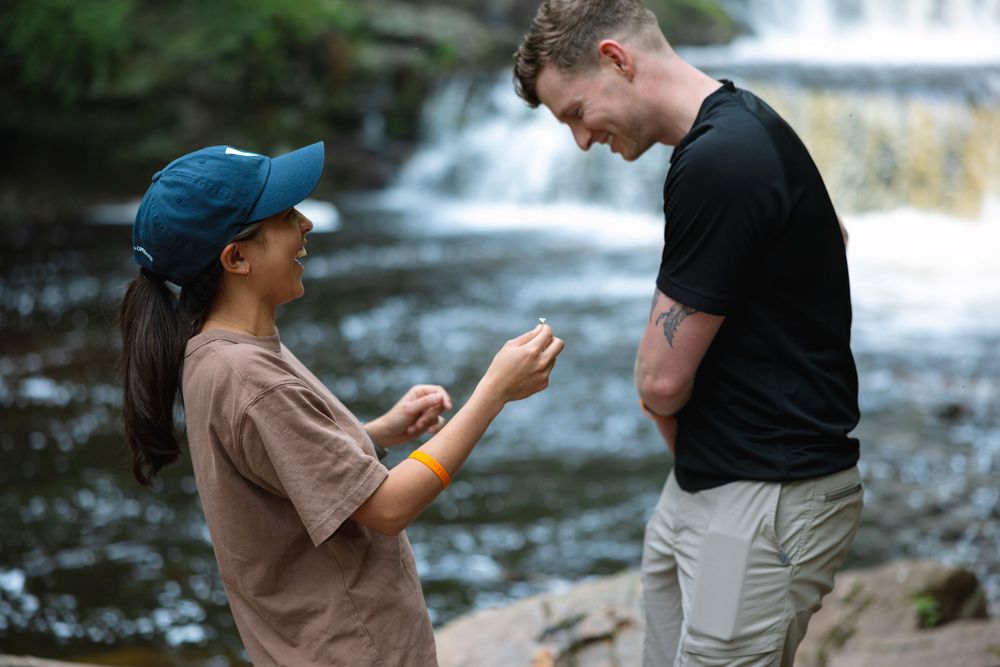 Couple examines engagement ring together while standing near a woodland waterfall.