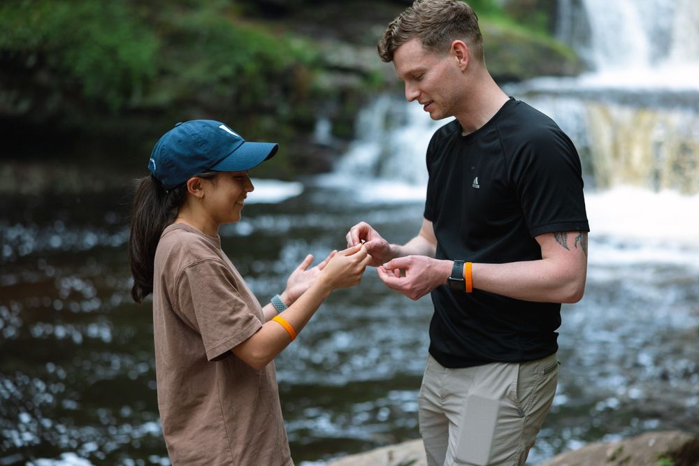 Man places engagement ring on womans finger beside a serene forest waterfall.