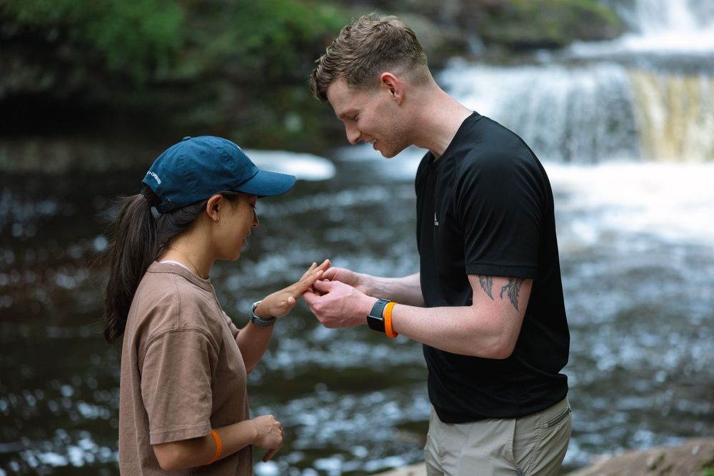 Close-up of man sliding engagement ring onto womans finger near a forest waterfall.