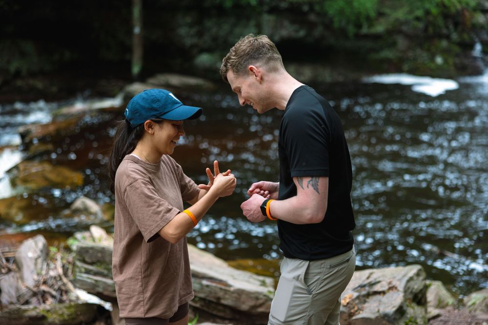 Man and woman sharing a moment by a river, looking at something small in their hands.
