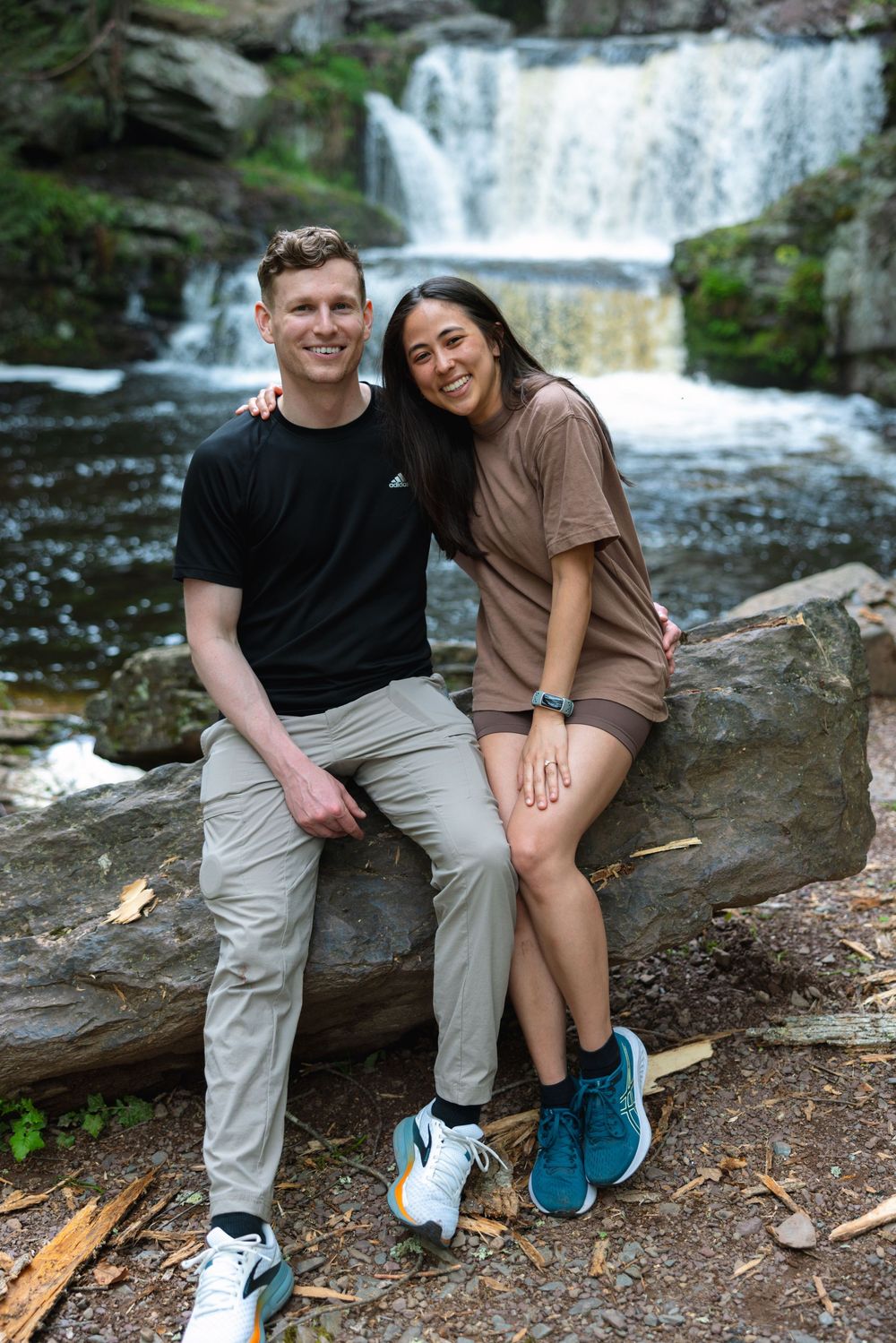 Smiling couple sitting on rocks in front of a beautiful waterfall in a lush forest.