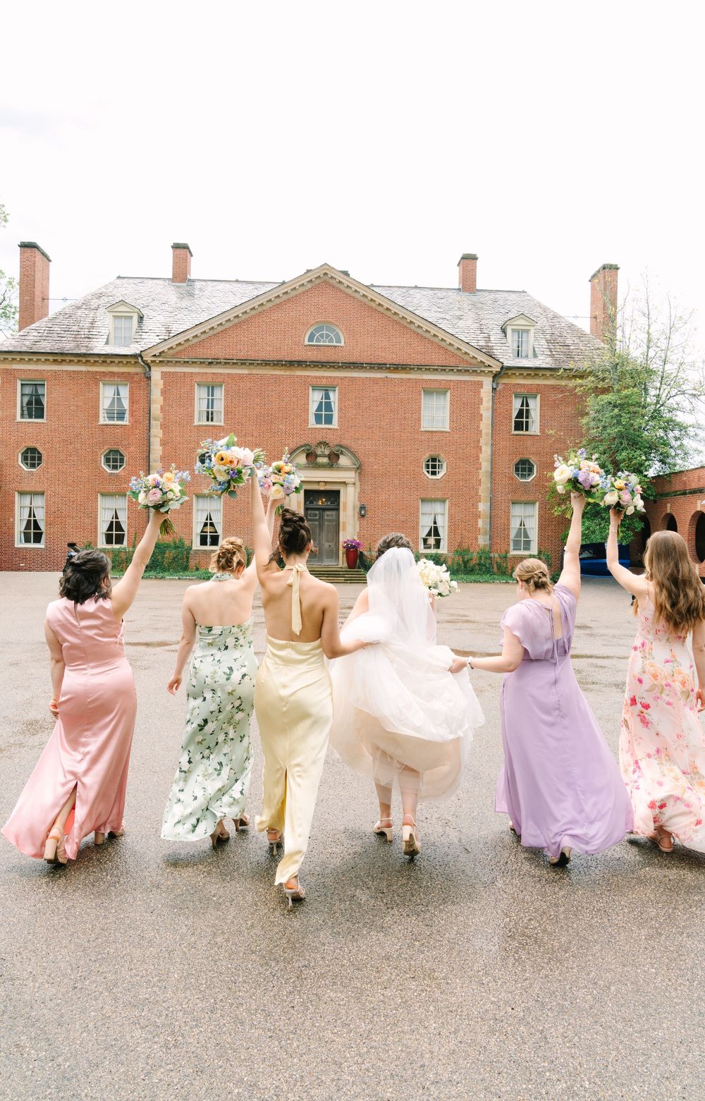 Bride and bridesmaids in colorful gowns raise bouquets walking toward elegant red brick mansion.
