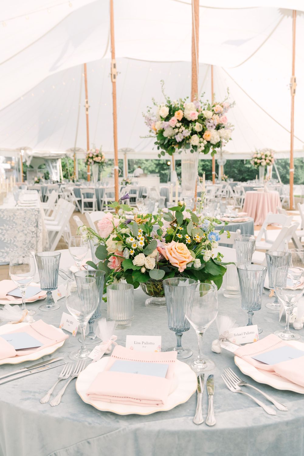 Elegant wedding reception table with gray linen, pink napkins, floral centerpieces, and blue goblets under a white tent.